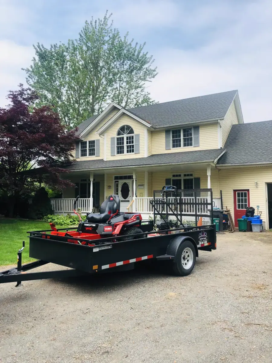 Trailer loaded with two red zero-turn mowers in front of a yellow house